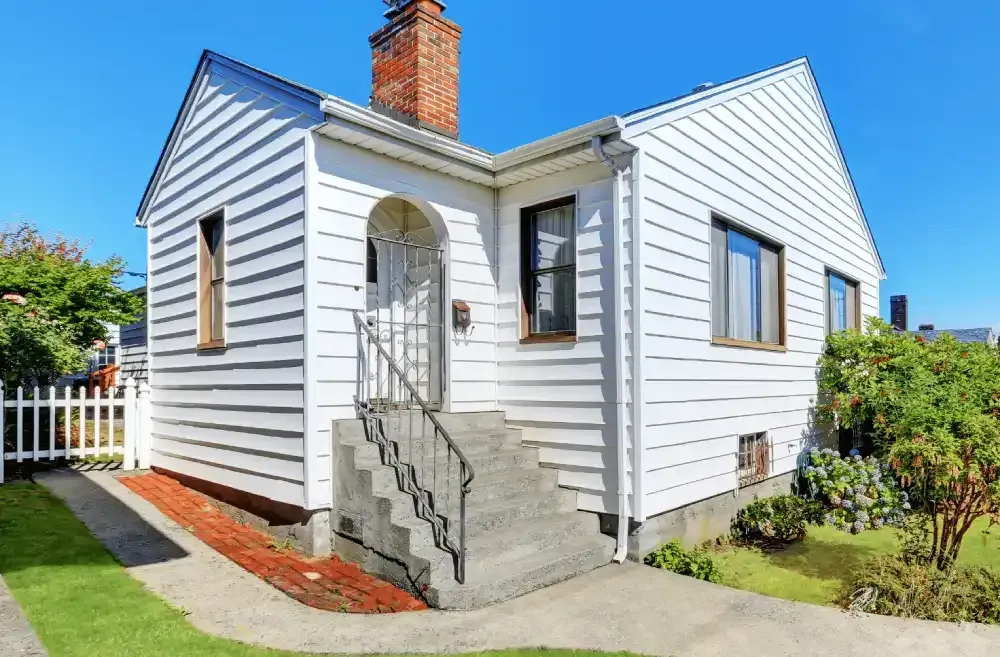 Small white house with red brick chimney, front steps, and a fenced yard in bright sunlight