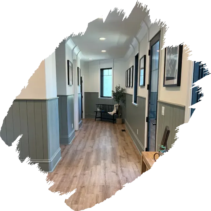 Long hallway with gray wainscoting, wood floors, framed art, and a small table by the far window.