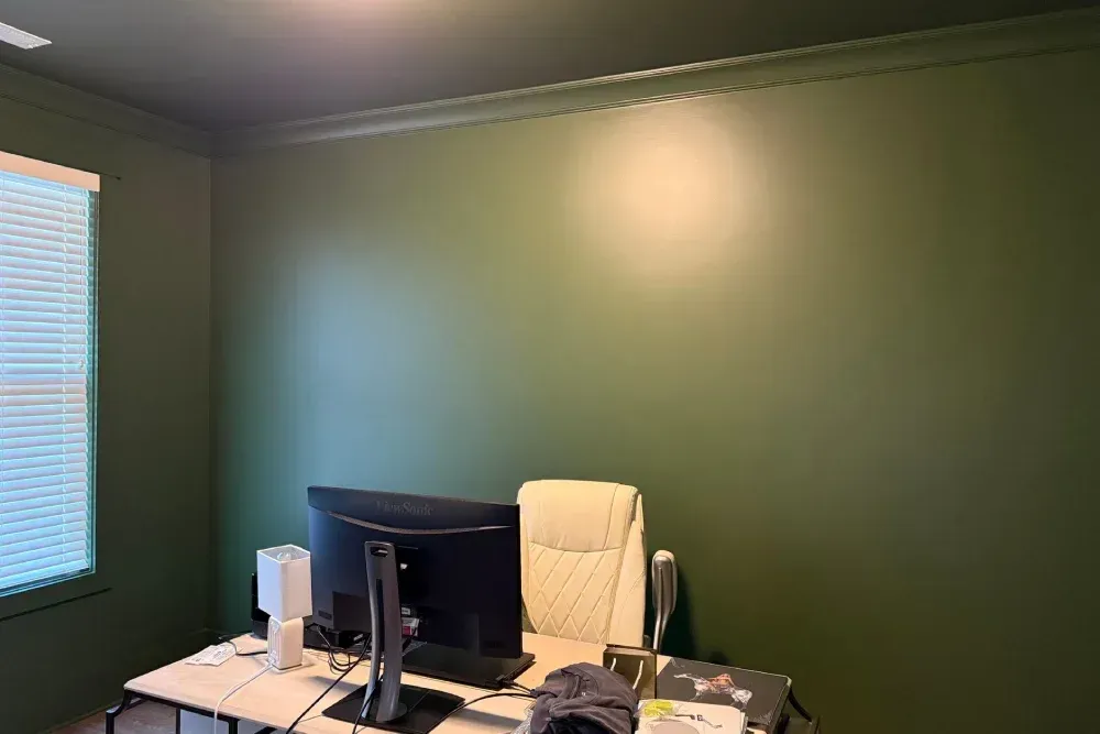 Empty home office with green walls, desk, monitor, and window blinds.