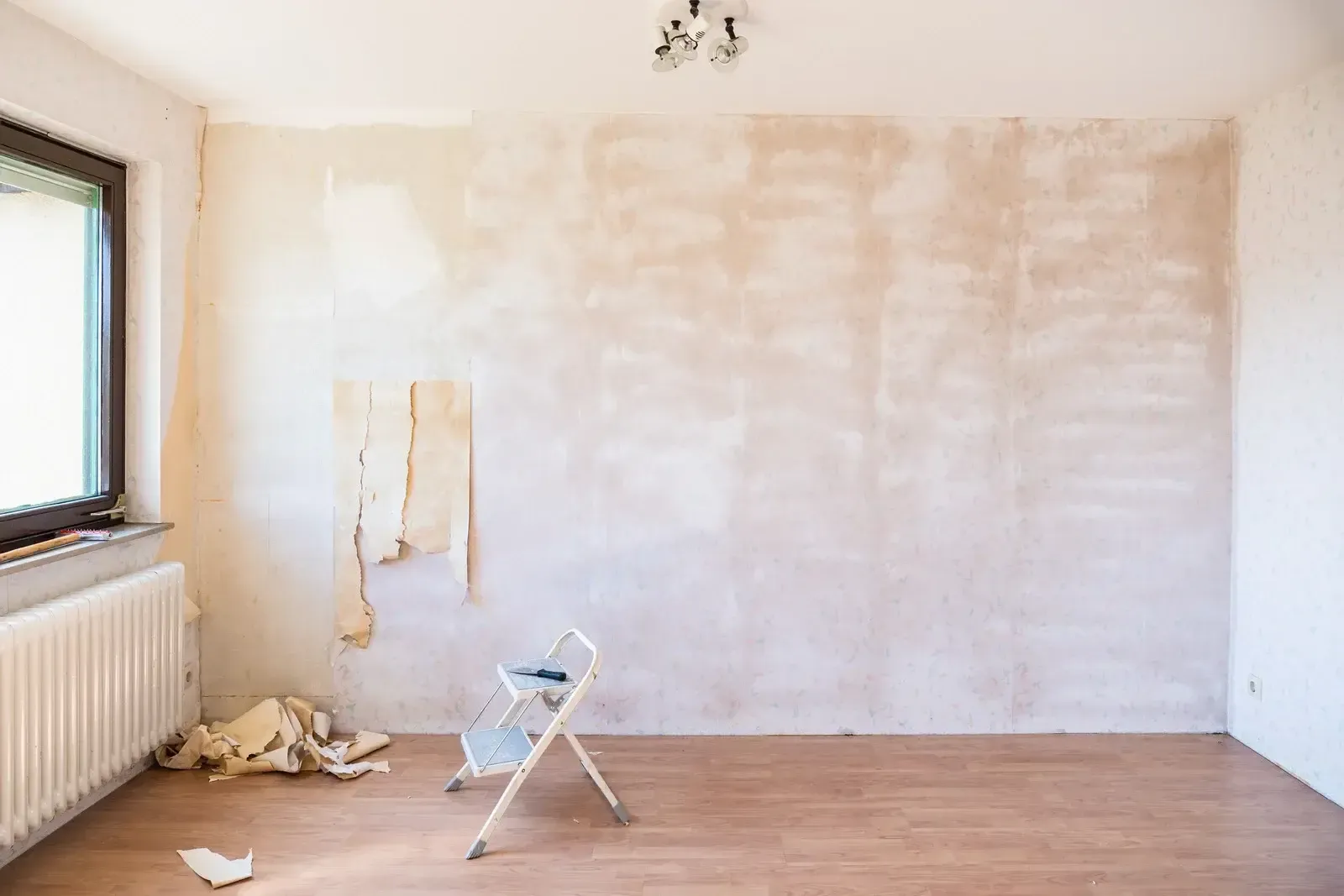 Sparse room with white walls, wooden floor, radiator, window, and a step stool in front of a partially painted wall