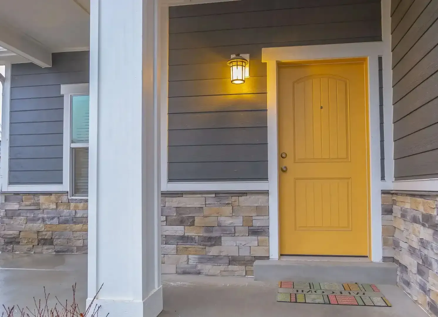 Front porch with a yellow door, wall lantern, gray siding, and stone accents on a house