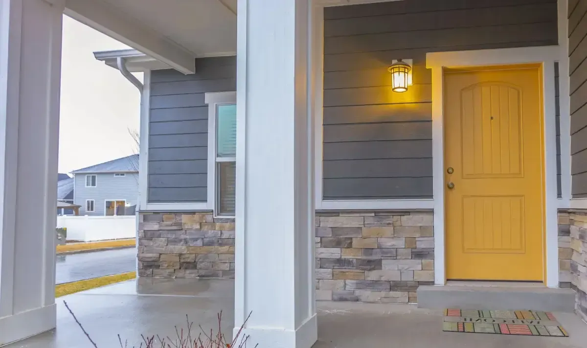 Front porch with gray siding, stone base, white pillars, and a bright yellow front door lit by a porch light.