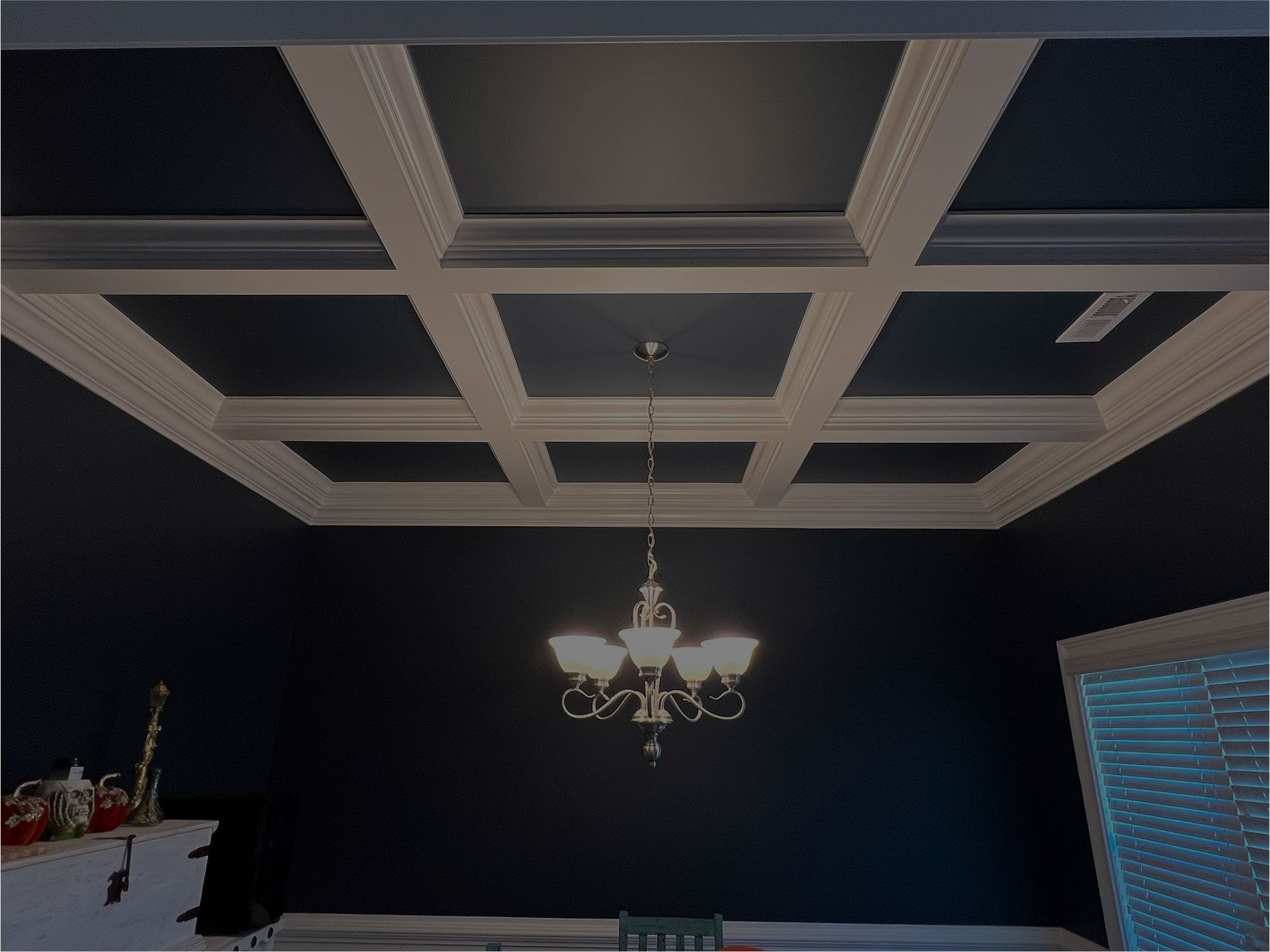 Dark dining room ceiling with white coffered panels, chandelier, and window blinds on the right.