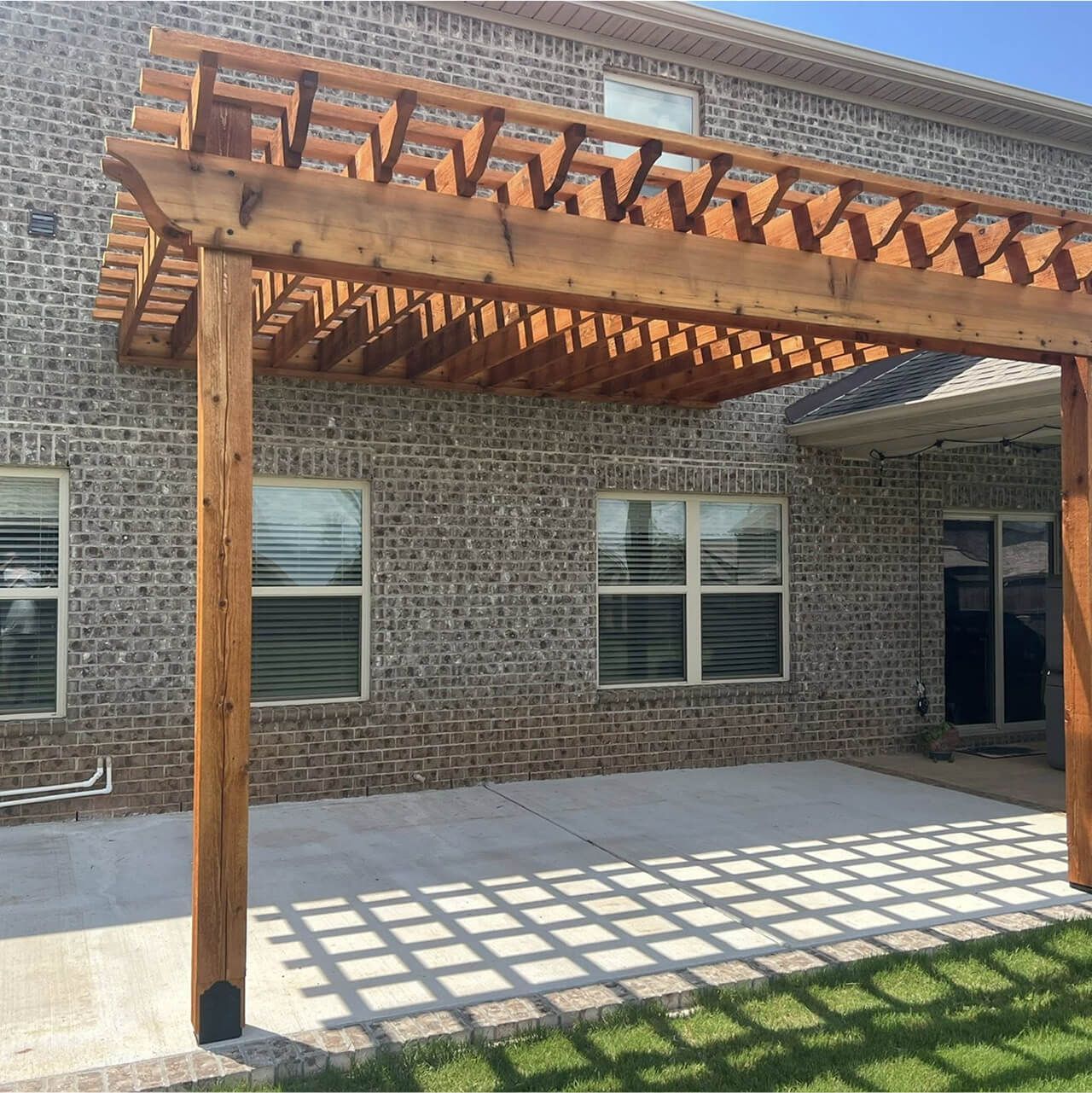 Wooden pergola patio beside a brick house, casting a lattice shadow on the concrete floor.