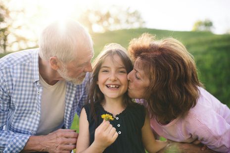 Smiling child between two adults outdoors, both leaning in to kiss her cheeks in warm sunlight