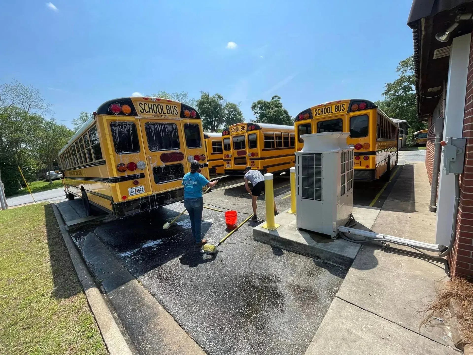 Two people washing school buses outside on a sunny day.