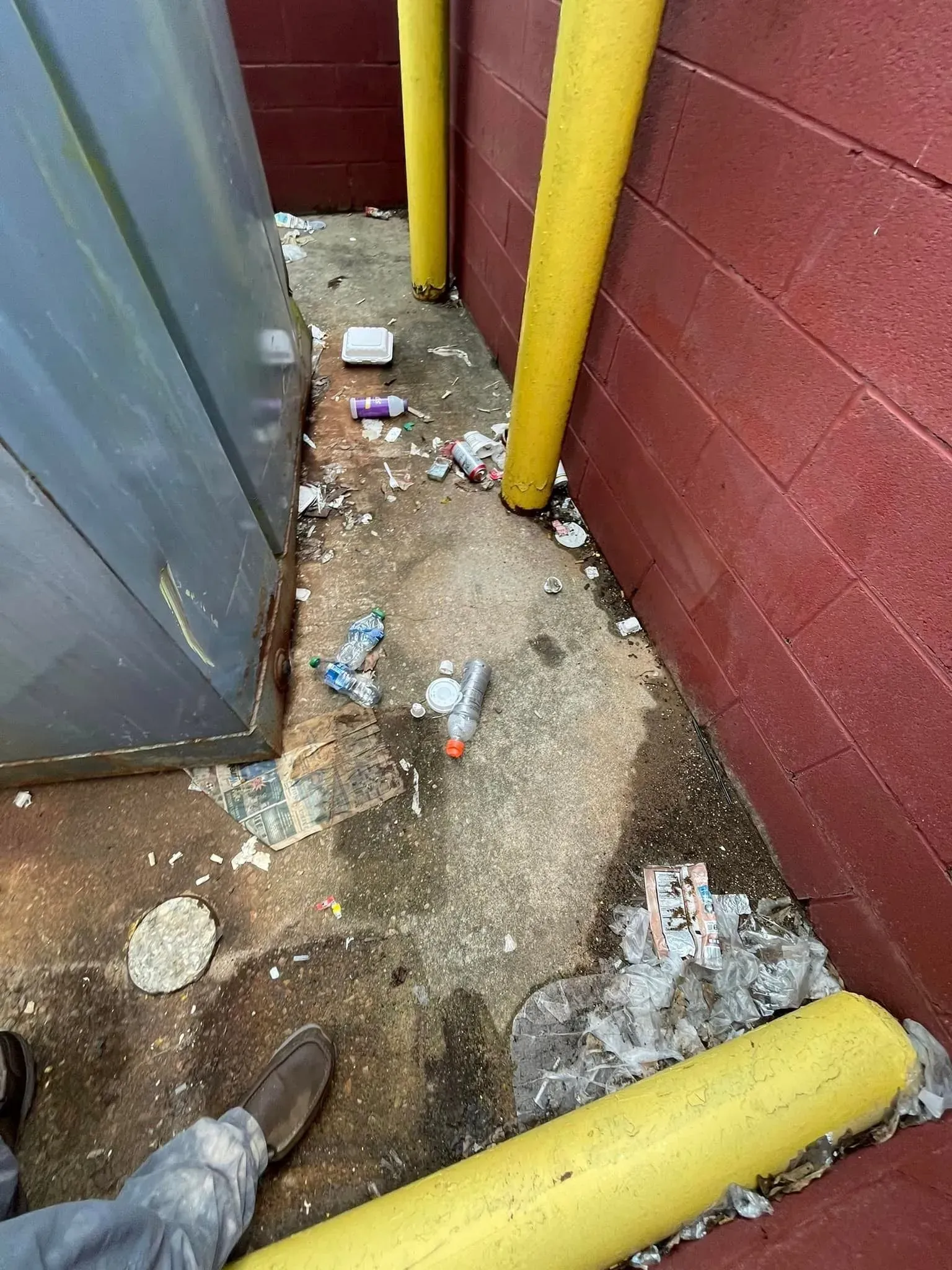 Trash-filled concrete area between red wall and metal container, with yellow safety poles.