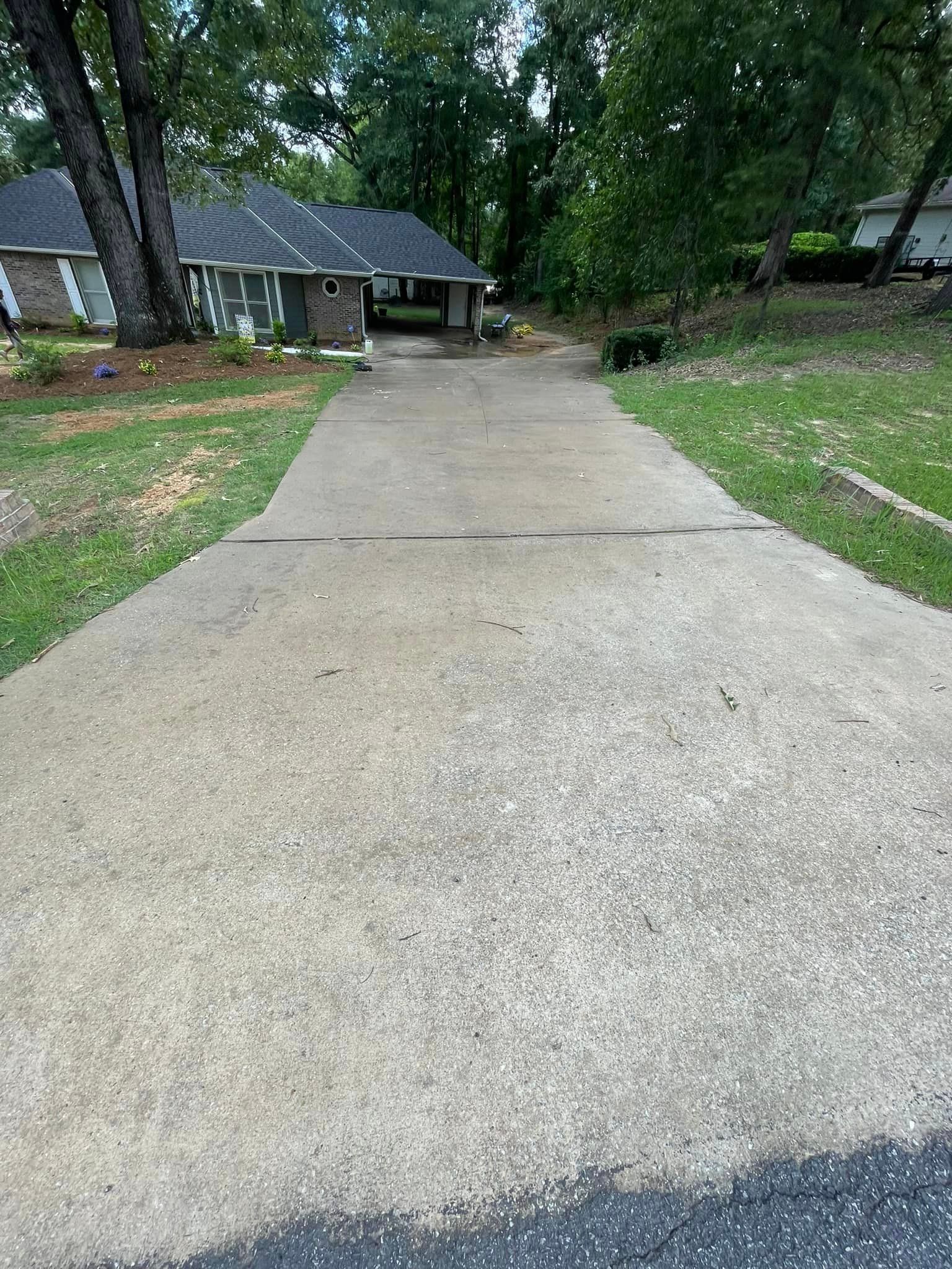 Concrete driveway leading to a house with a dark roof, surrounded by grass and trees.