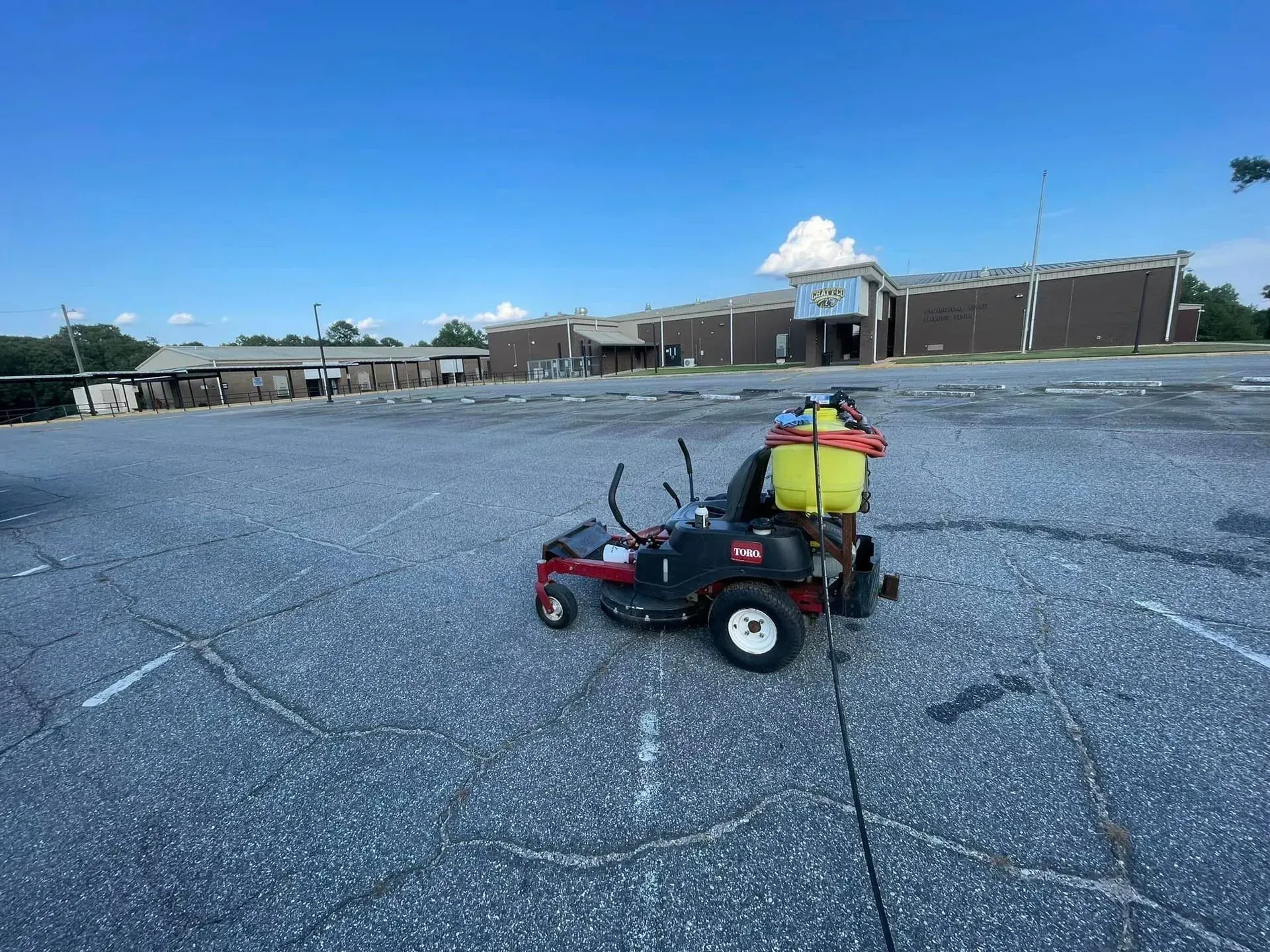 Zero-turn mower with yellow sprayer in a parking lot, building in the background, blue sky.