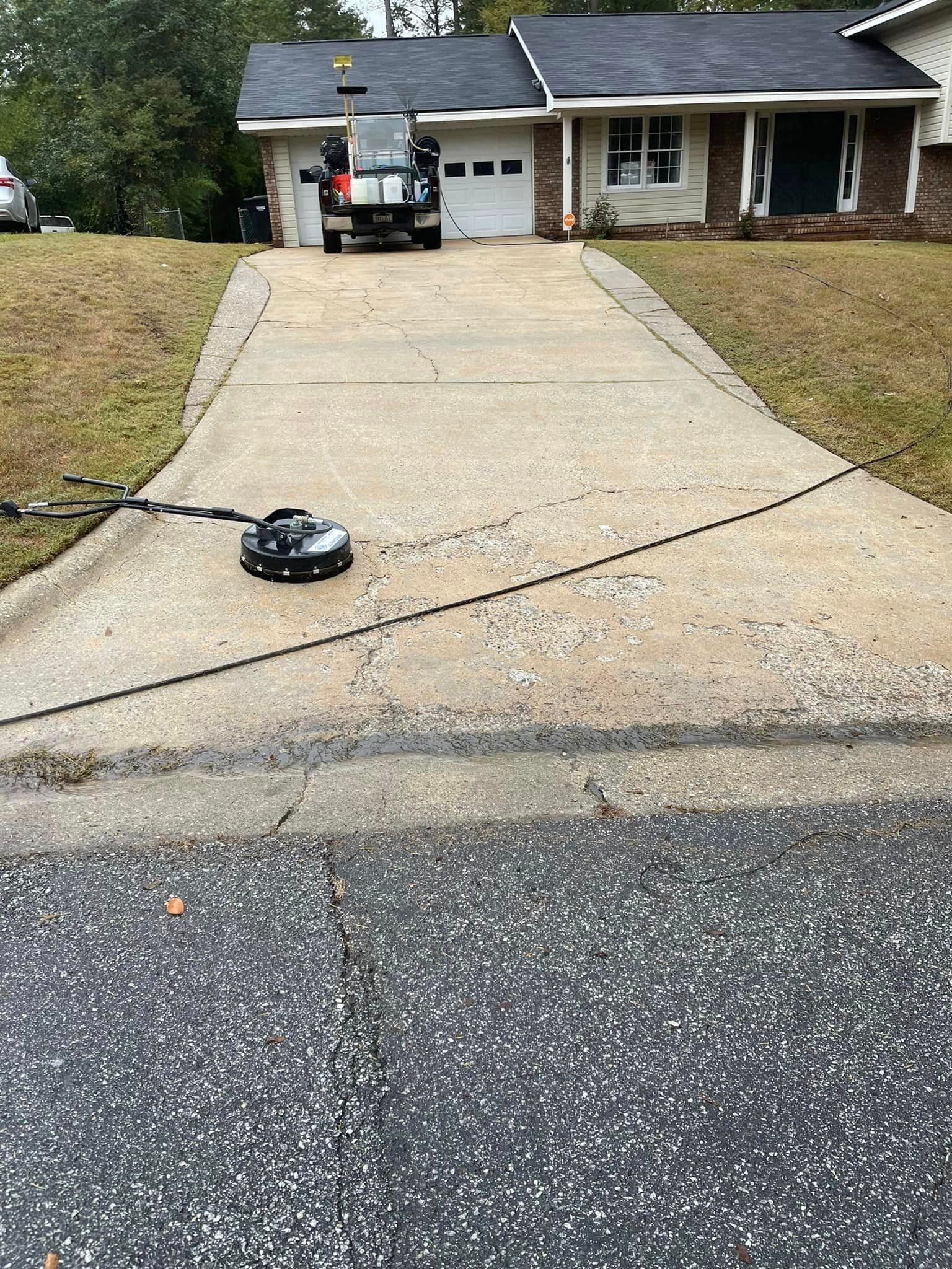 A black pressure washer on a concrete driveway in front of a house. A truck is parked in the garage.