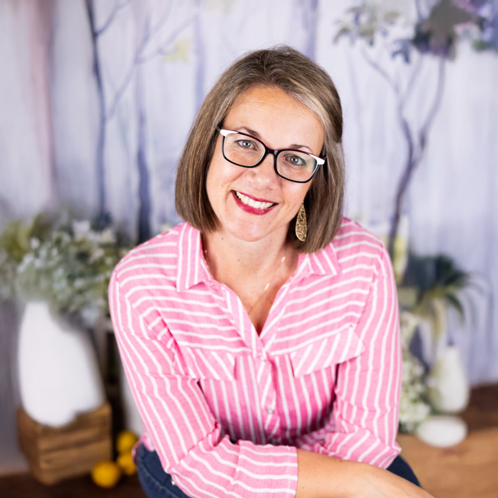 Smiling woman in glasses wearing a pink striped shirt, seated indoors with a soft floral backdrop