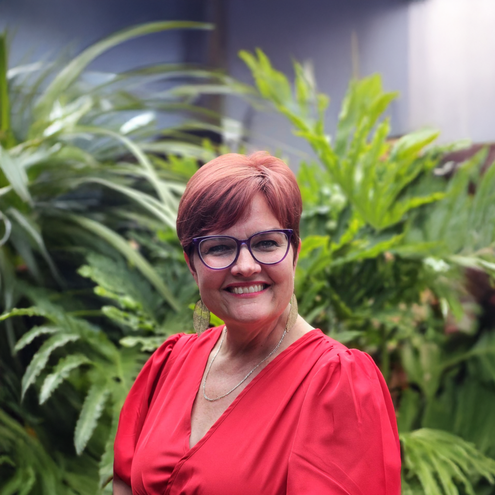 Smiling person in red blouse and glasses standing in front of lush green plants