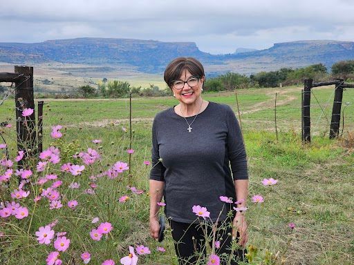 Woman standing in a flower field with mountains in the background, smiling and wearing a dark top.
