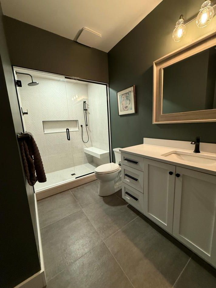 Small modern bathroom with white vanity, mirror, toilet, and glass shower on dark wood flooring