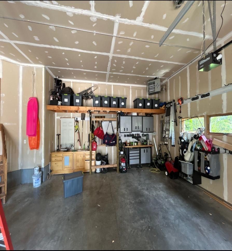 Cluttered garage with storage shelves, workbench, tools, and hanging gear under unfinished drywall.