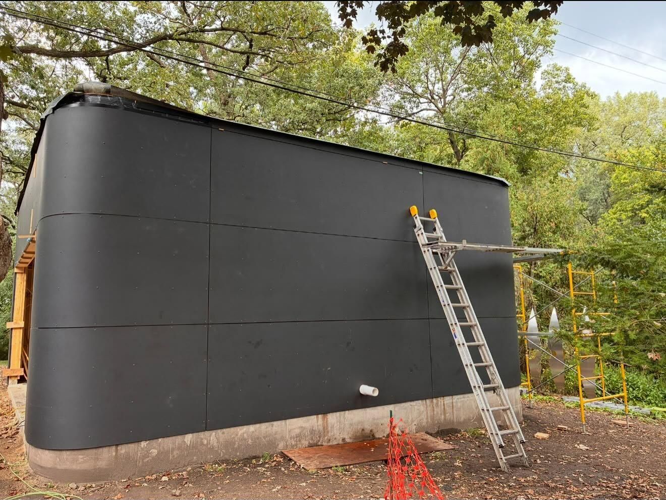 Large black cylindrical tank outdoors, with a ladder leaning against it near trees.