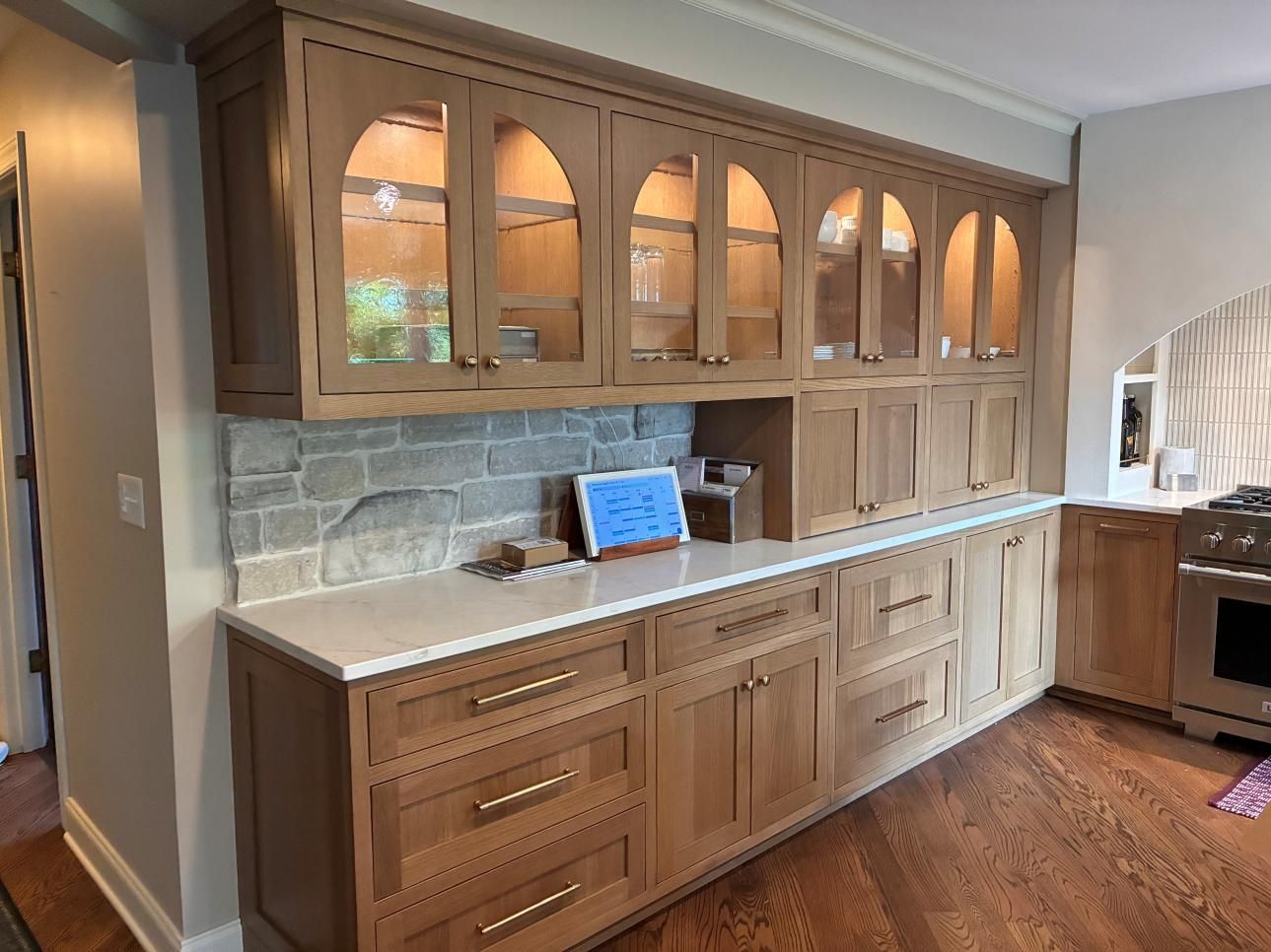 Wooden kitchen hutch with glass-front cabinets, stone backsplash, and countertop in a bright room