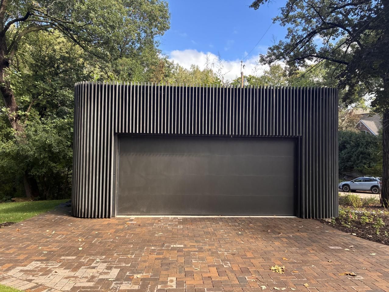 Modern black garage with vertical slats, set behind a brick driveway and trees.