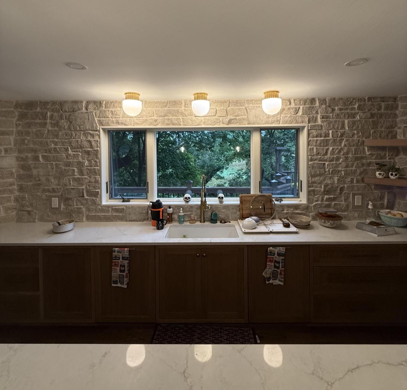 Brick kitchen with wide window over sink, white countertop, dark cabinets, and three pendant lights.