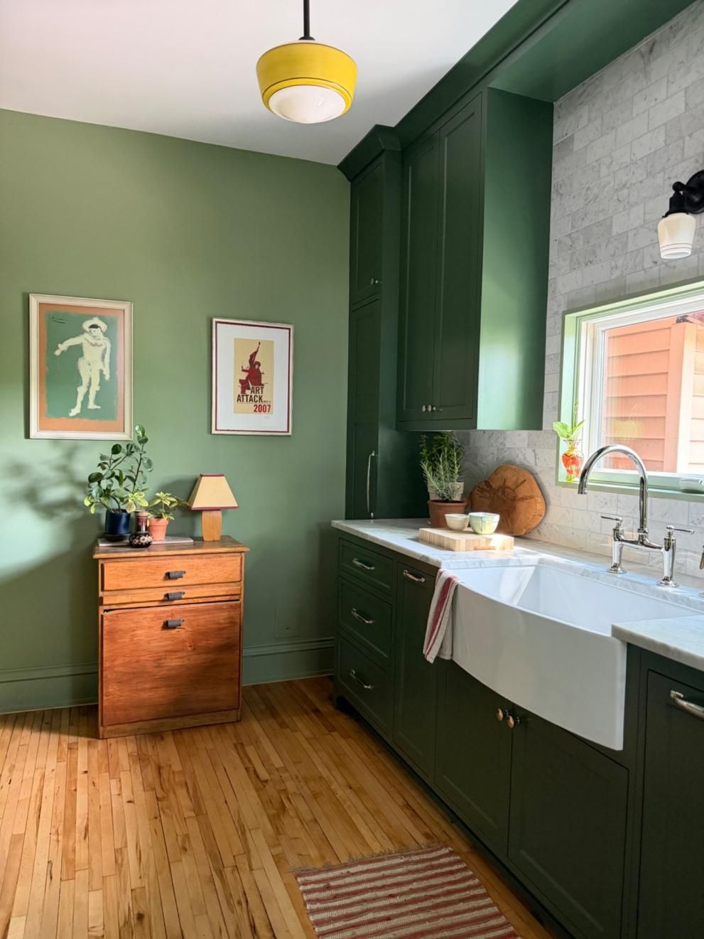 Green kitchen with dark cabinets, white sink, wood floors, and a small dresser beside framed wall art