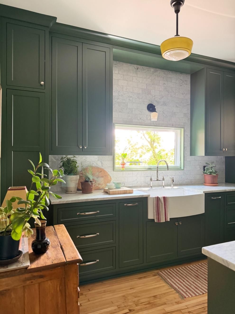 Dark green kitchen with farmhouse sink, window above counter, wood accents, and potted plants.