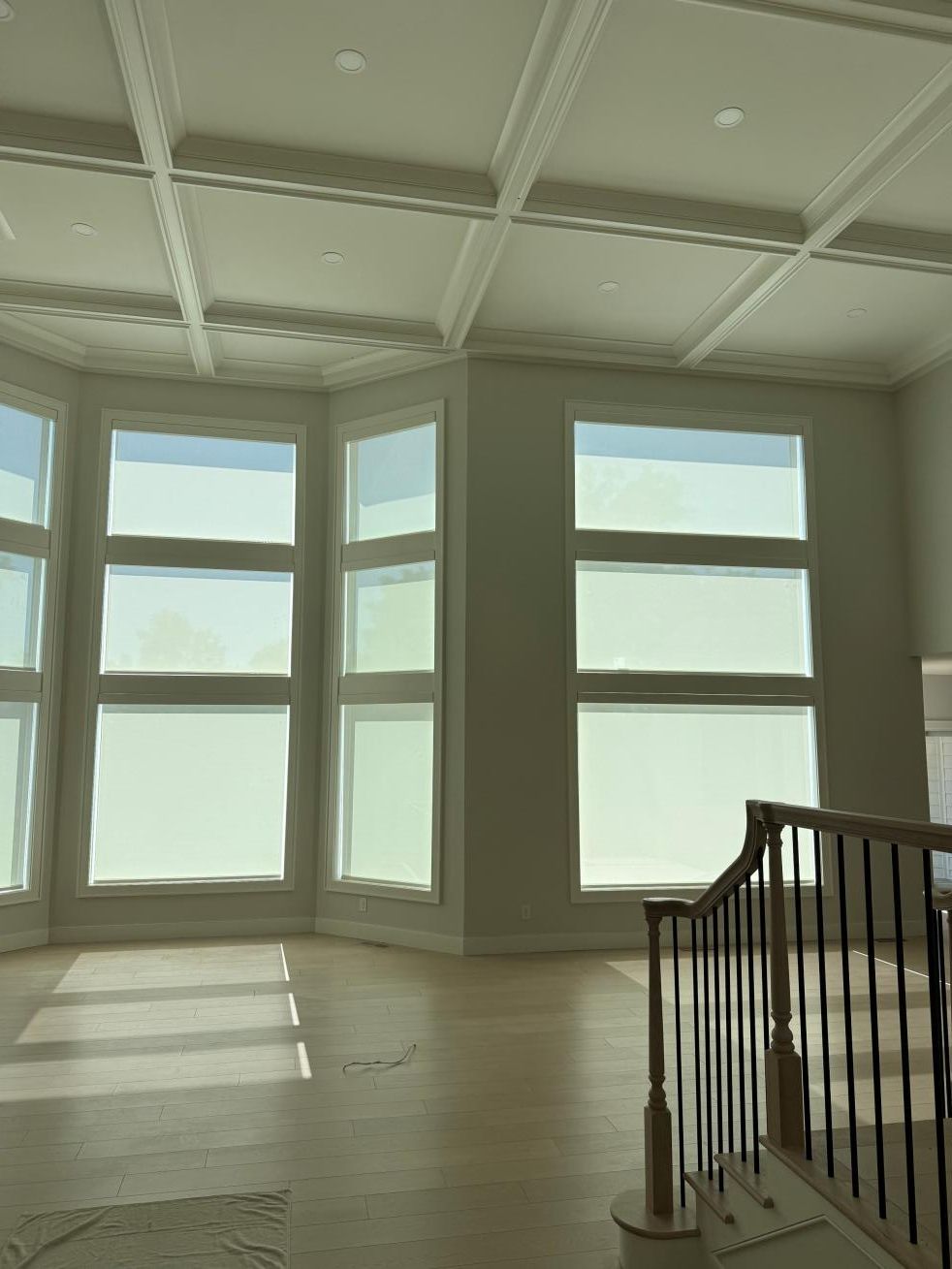 Bright empty room with large bay windows, sunlight on hardwood floors, and a staircase railing on the right