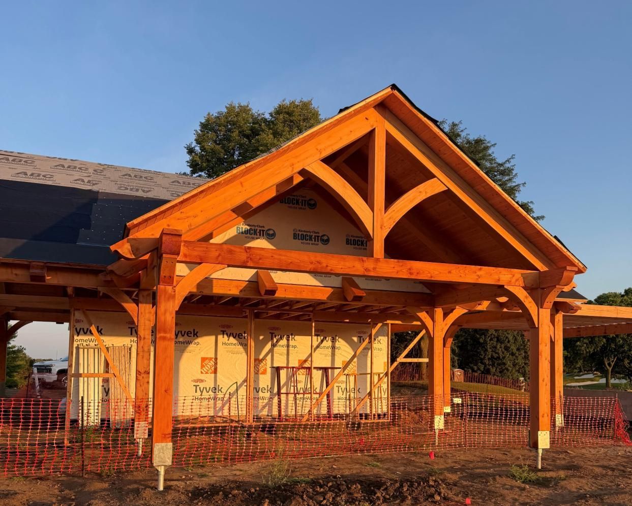 Sunlit wooden pavilion with exposed beams and a gabled roof under construction on a grassy lot