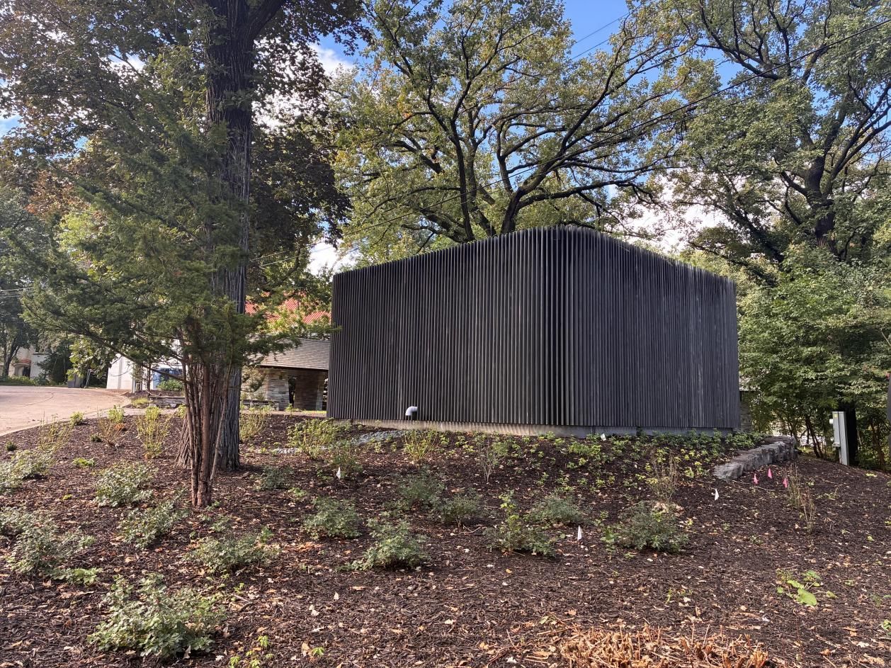 Black corrugated metal shed among trees on a wooded hillside, with a white pipe visible at the base