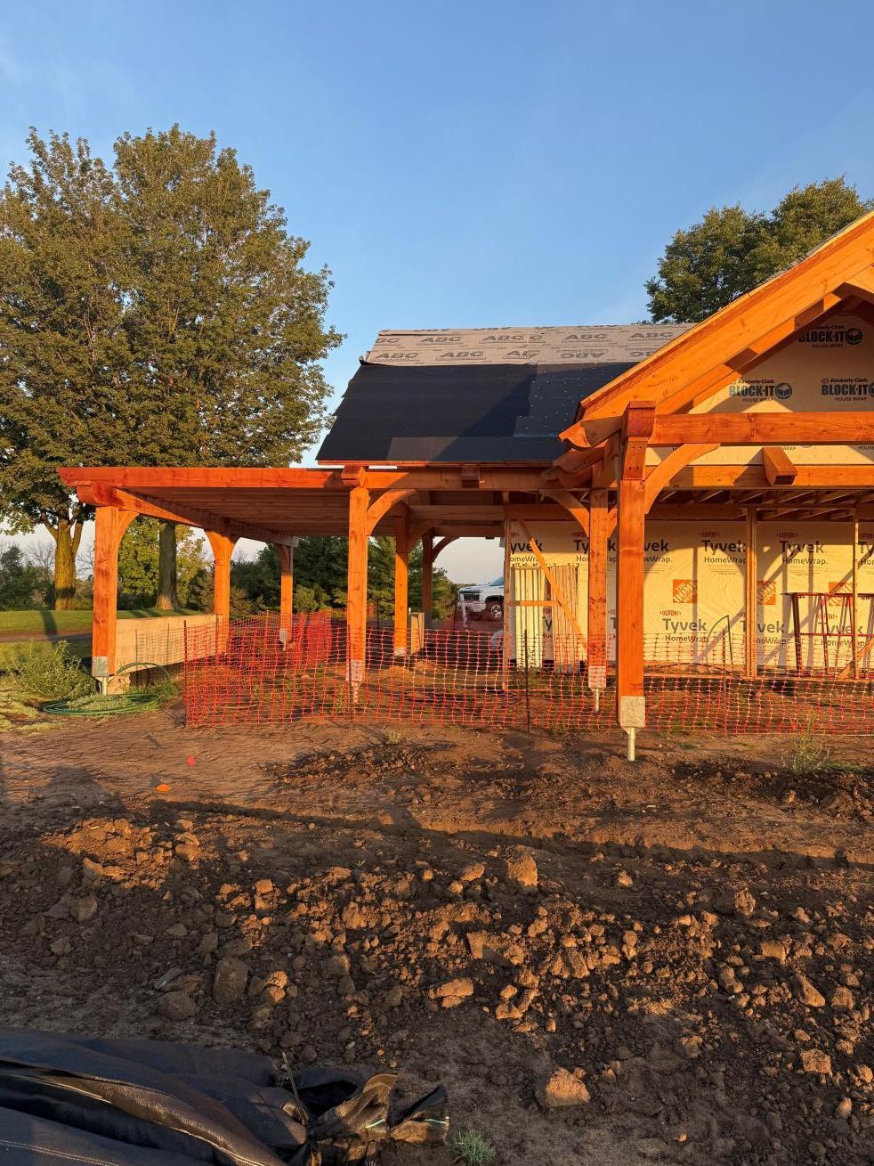 Orange wooden pavilion under construction beside a stone wall, with trees and blue sky in the background