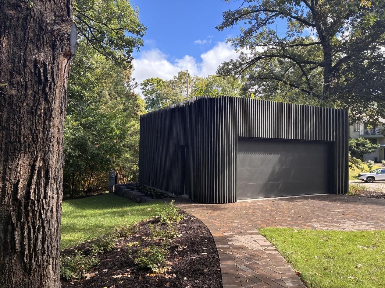 Dark modern garage/shed with black vertical siding beside a paved path in a wooded yard