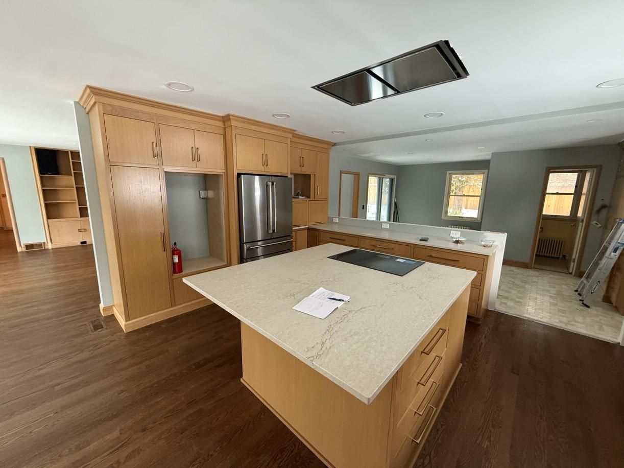 Empty modern kitchen with wood floors, island, stainless fridge, and recessed ceiling opening.