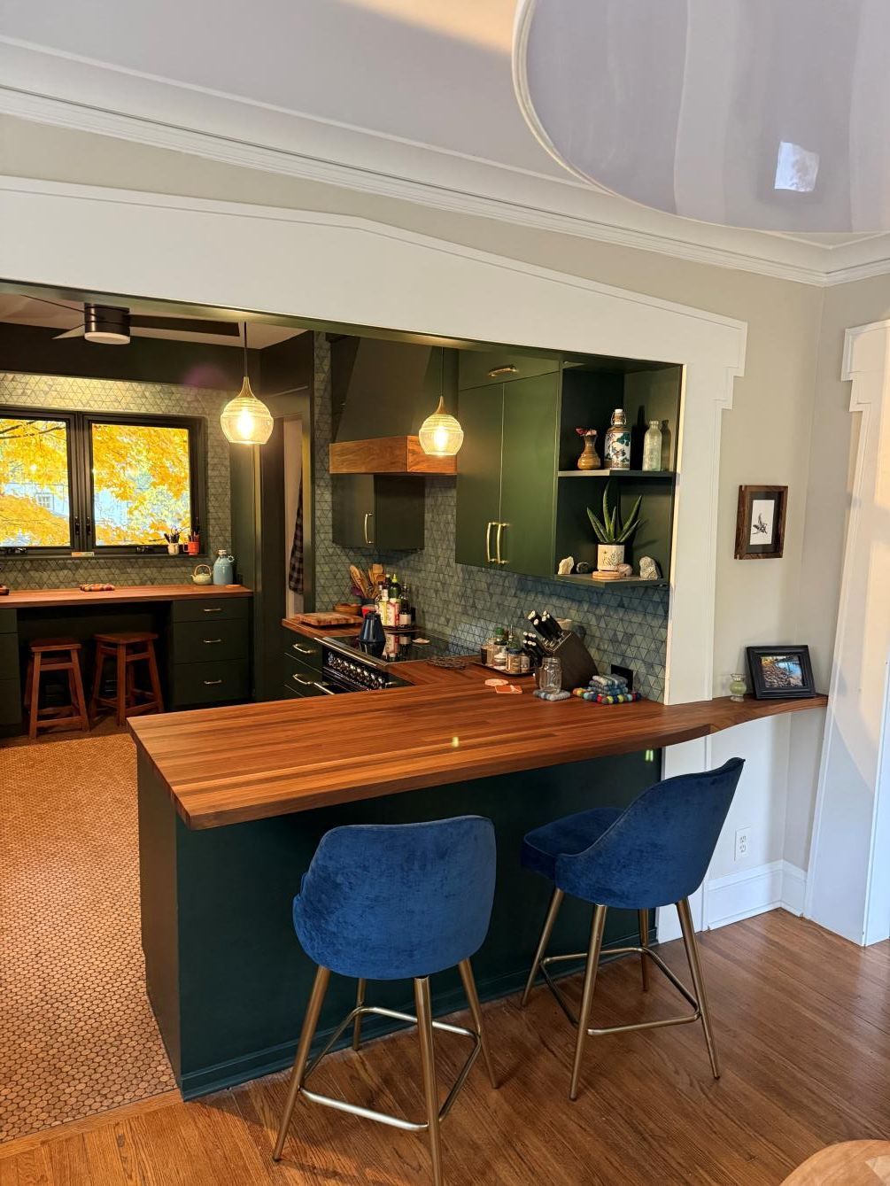 Home bar with wood counter, two blue stools, and a dark green kitchen opening behind it