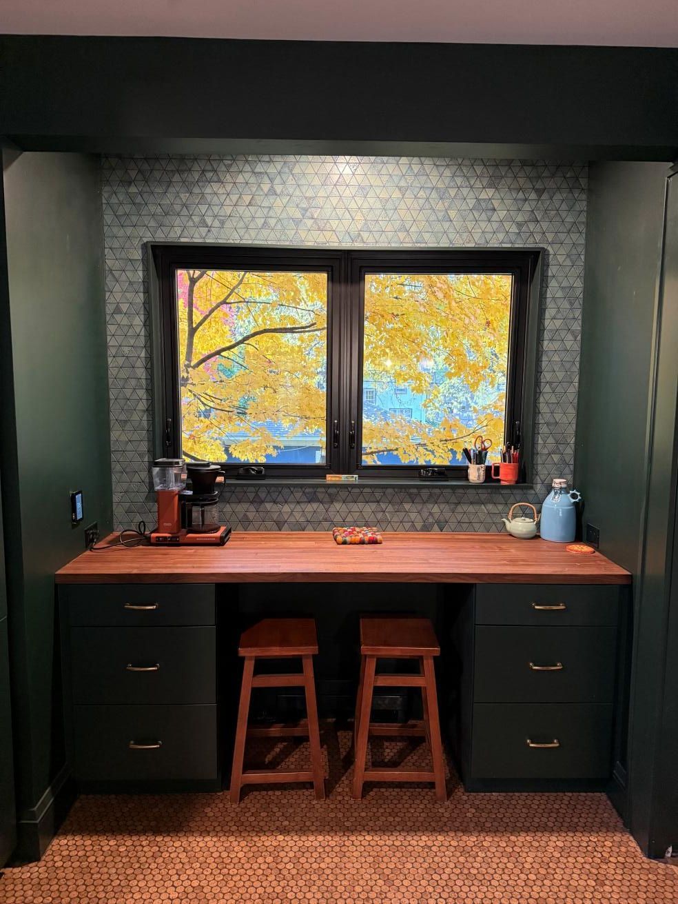 Built-in desk nook with dark cabinetry, wood countertop, two stools, and window view of autumn trees.