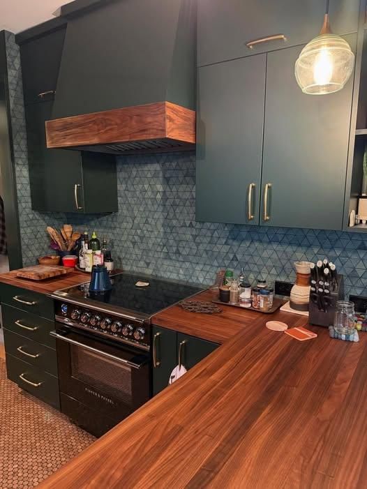 Modern kitchen with dark cabinets, blue tile backsplash, wood countertops, and a stove under a hood.