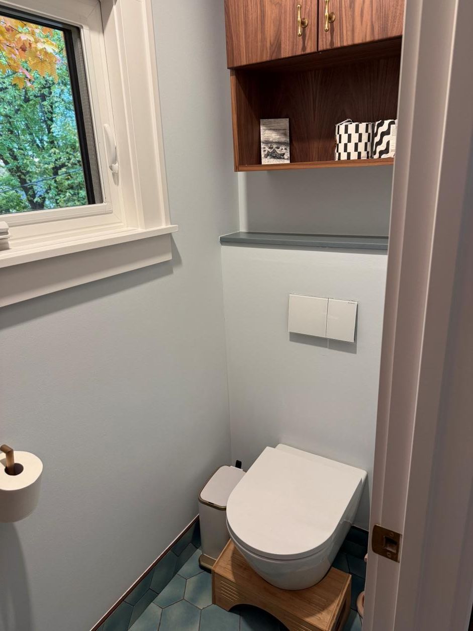 Small bathroom with a toilet under a shelf, gray walls, and a window on the left.