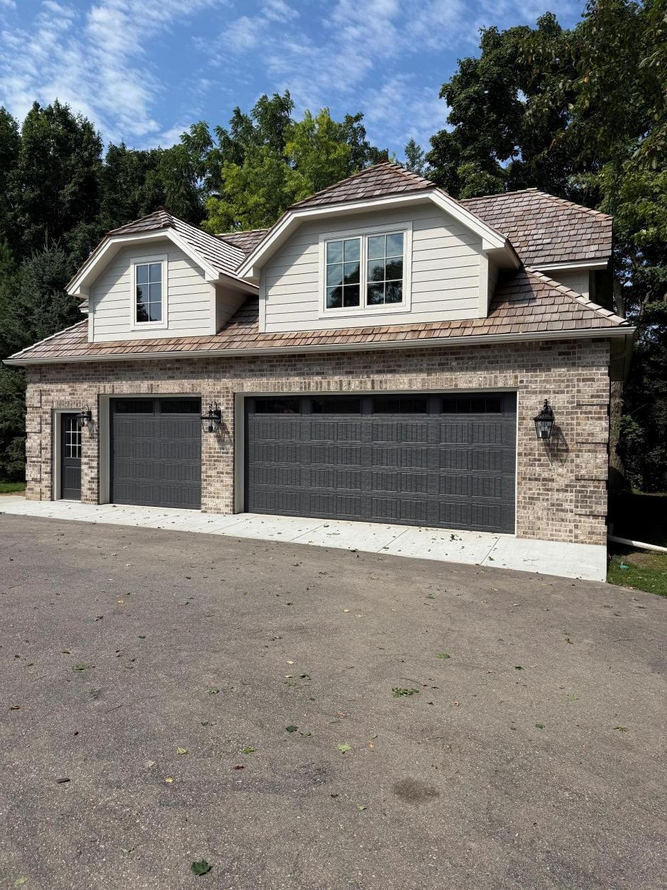 Stone two-car garage with dark doors and dormer windows under a wooded sky