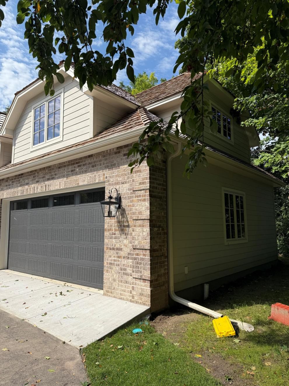 Suburban two-story house with stone facade, gray garage door, and trees casting shade over the driveway.