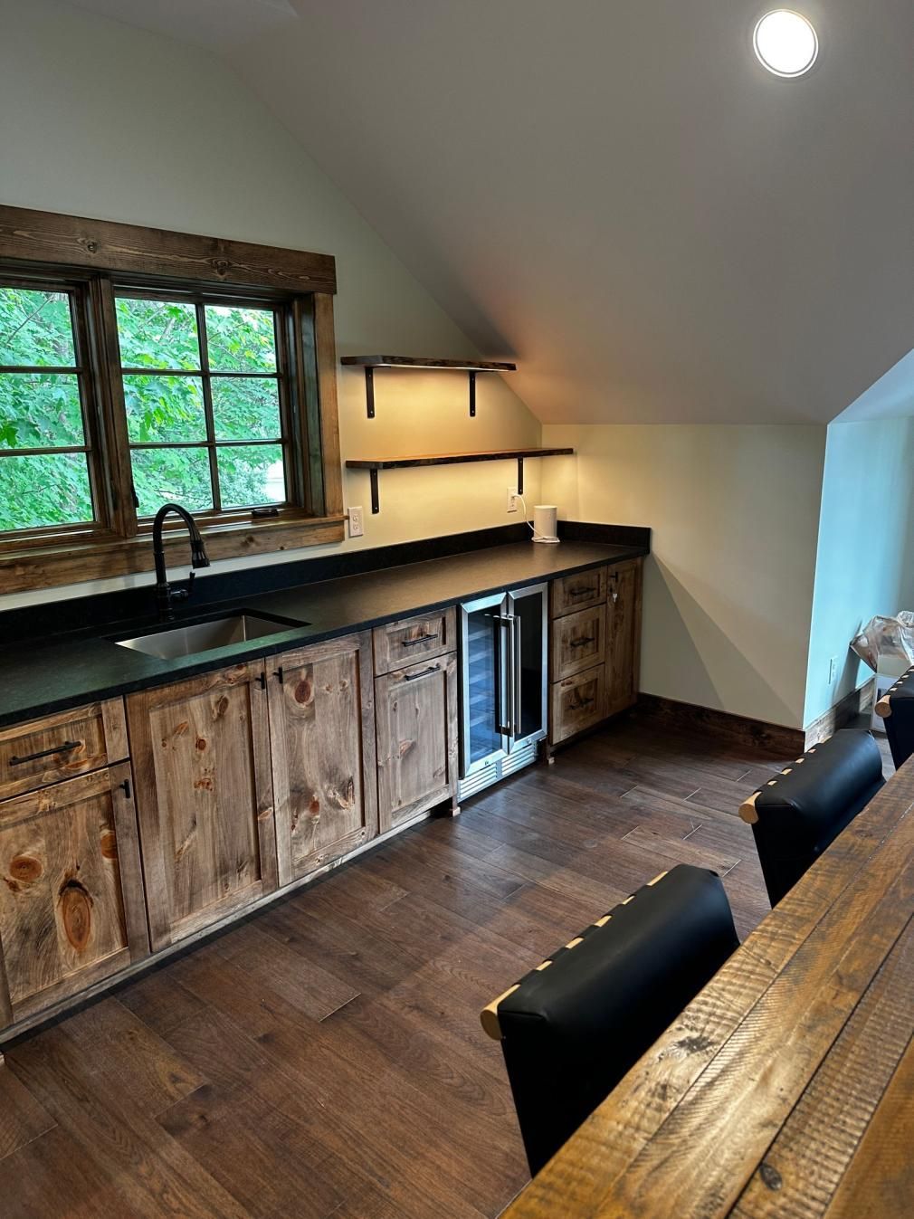 Rustic kitchen with wooden cabinets, black countertops, window, and breakfast bar under sloped ceiling