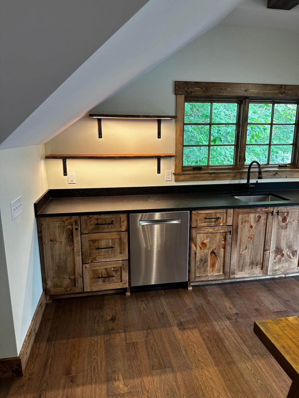 Small rustic kitchen nook with wood cabinets, stainless dishwasher, open shelves, and a window above the sink.