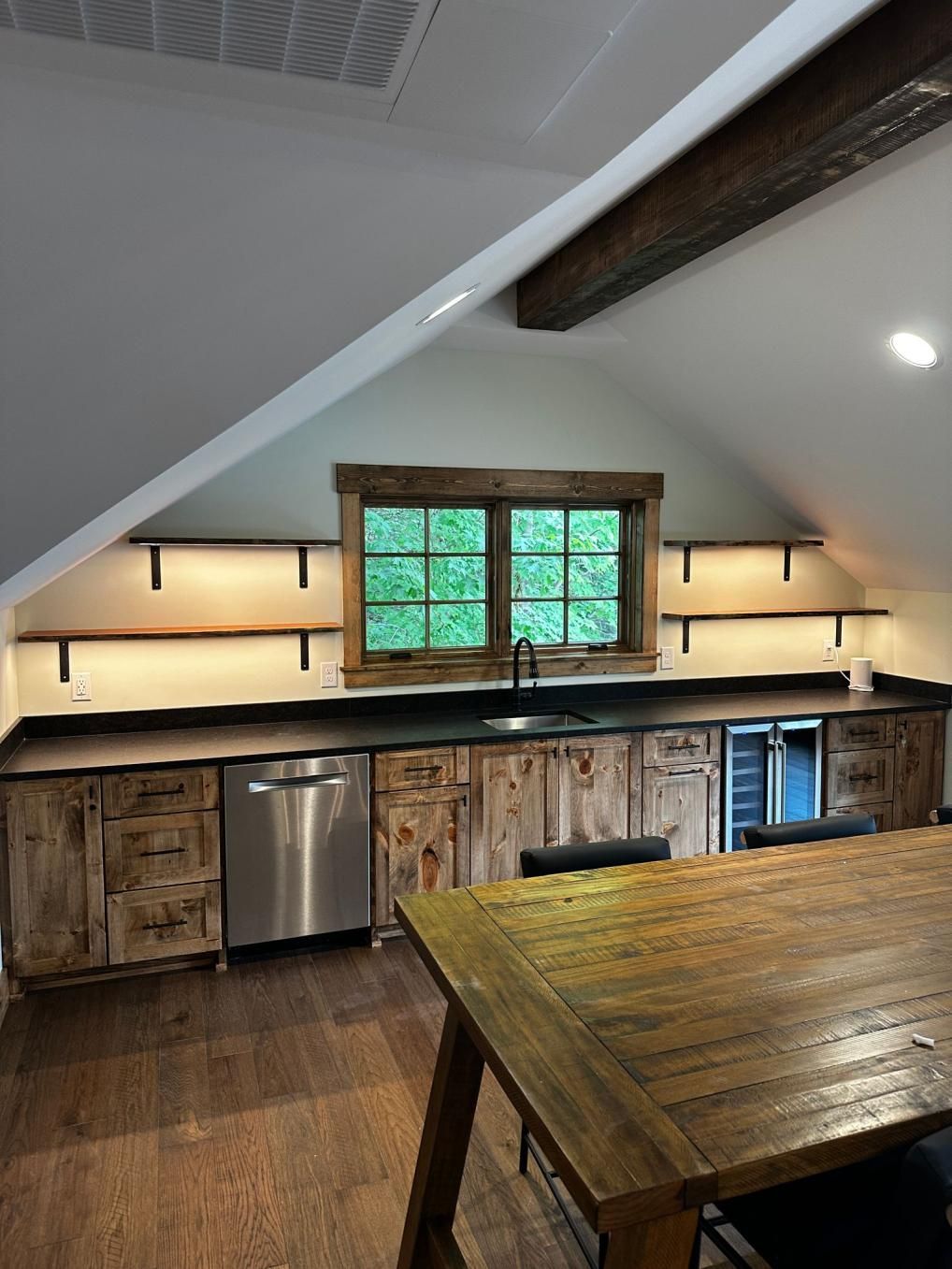 Rustic attic kitchen with wood table, open shelves, stainless fridge, and a small window over the counter