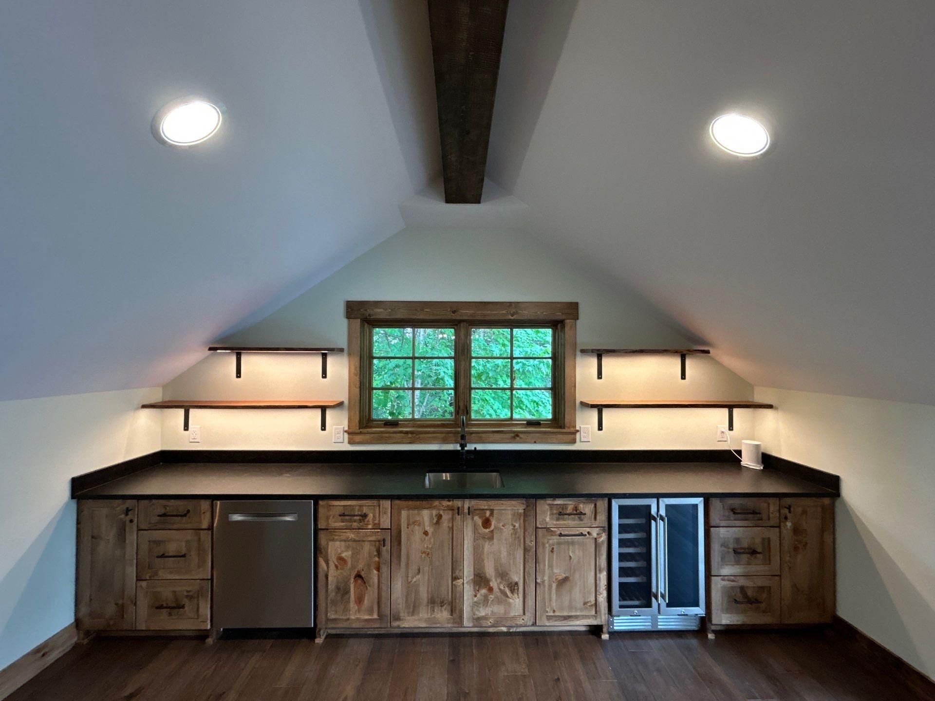 Attic kitchenette with rustic wood cabinets, black countertops, and a small window under sloped white ceiling