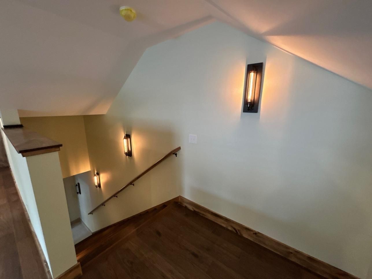 Stairwell landing with white walls, wooden steps, and wall sconces in a narrow attic-like space