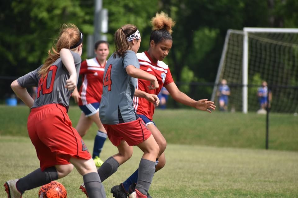 Soccer players in red and gray uniforms chase the ball on a grassy field near the goal