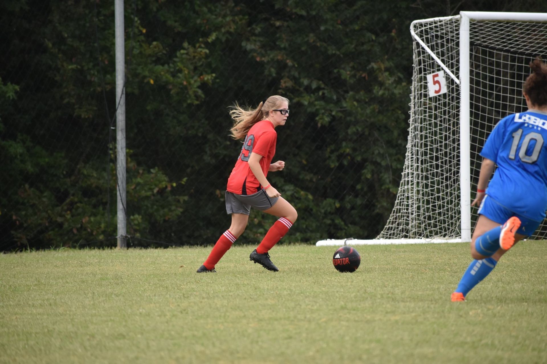 Soccer player in red dribbles toward goal while a blue defender approaches on the field