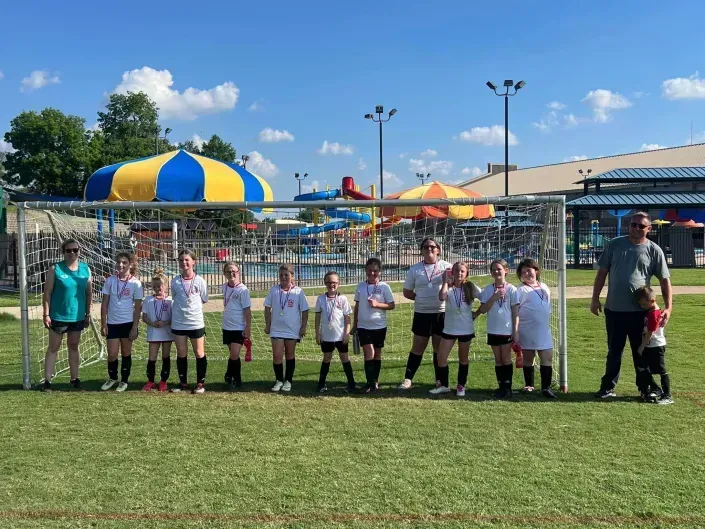 Youth soccer team lined up on a field with coaches, in front of colorful tents and playground equipment.