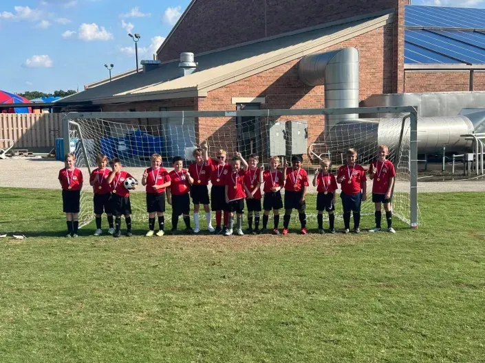 Youth soccer team lined up on a grassy field in front of a brick building and goal net.