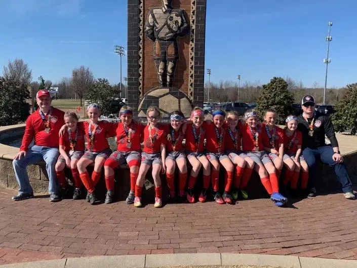 Group in red sports uniforms seated on a bench in front of a tall statue outdoors.