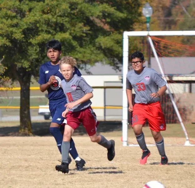 Two soccer players chase the ball on a sunny field, with a goal visible in the background.