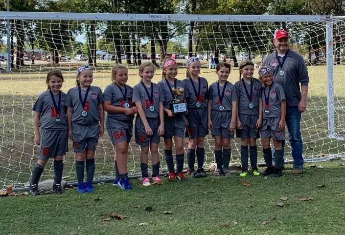 Youth soccer team posing in front of a goal on a grassy field, wearing dark uniforms and medals.