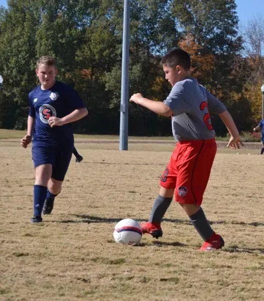 Two soccer players contesting the ball on a grassy field, one in gray and red, one in navy blue.